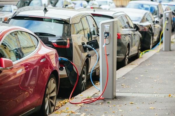 A row of electric cars parked on a street, with charging leads trailing over the pavements to the lamposts.