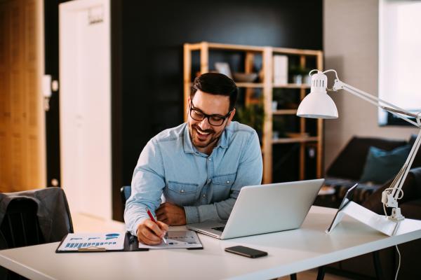 Man working at desk in a home office with laptop and writing in a clipboard