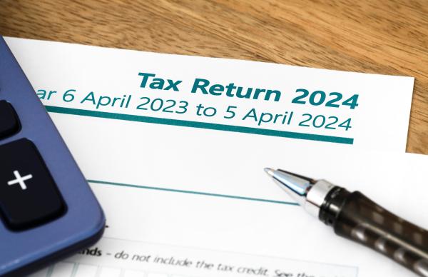 Paper income tax return for tax year 2023-24 on wooden table, with calculator and pen laid on top.