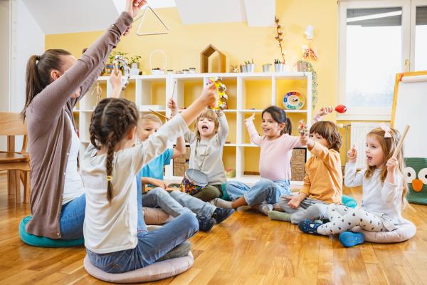 Nursery scene - children sat on the floor with arms raised copying a nursery teacher