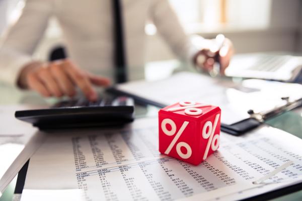 In the background, a man in a white shirt and black tie sits at a desk with a clipboard and calculator.  In the foreground sit sheets of figures, with a red dice like cube with % signs on it in white.