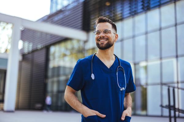 Male nurse stood outside a glass building wearing blue medical scrubs