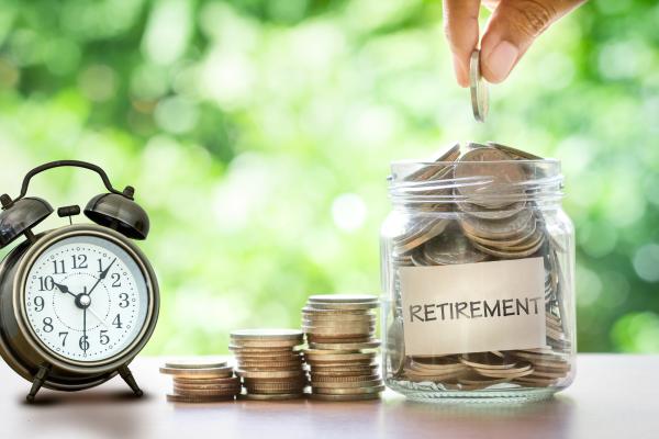 Hand putting coins from a pile into a jar labelled retirement, with an alarm clock on the left of the image